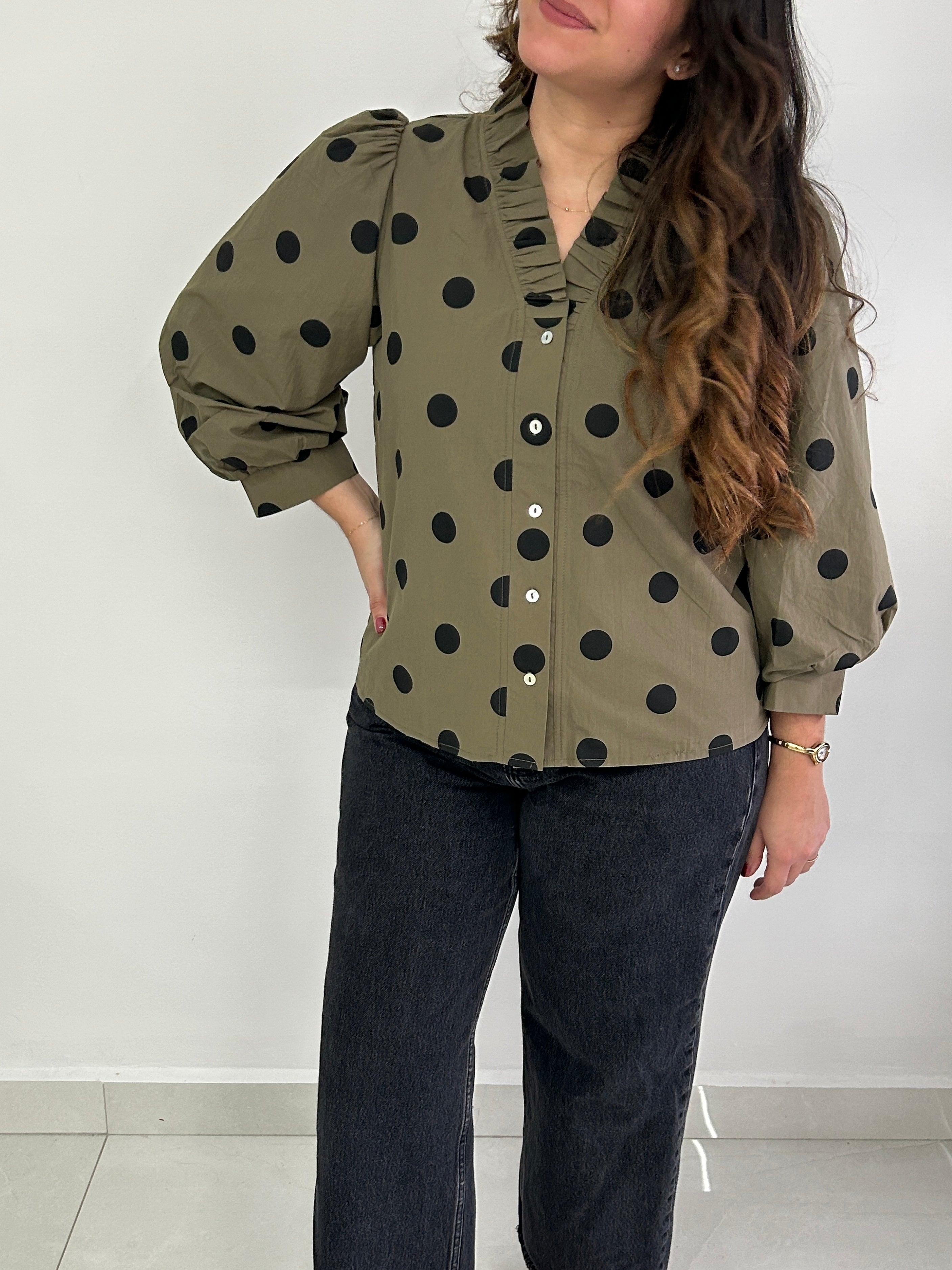 A woman wearing an olive green long-sleeve blouse with black polka dots, paired with dark jeans, standing against a plain light gray wall.