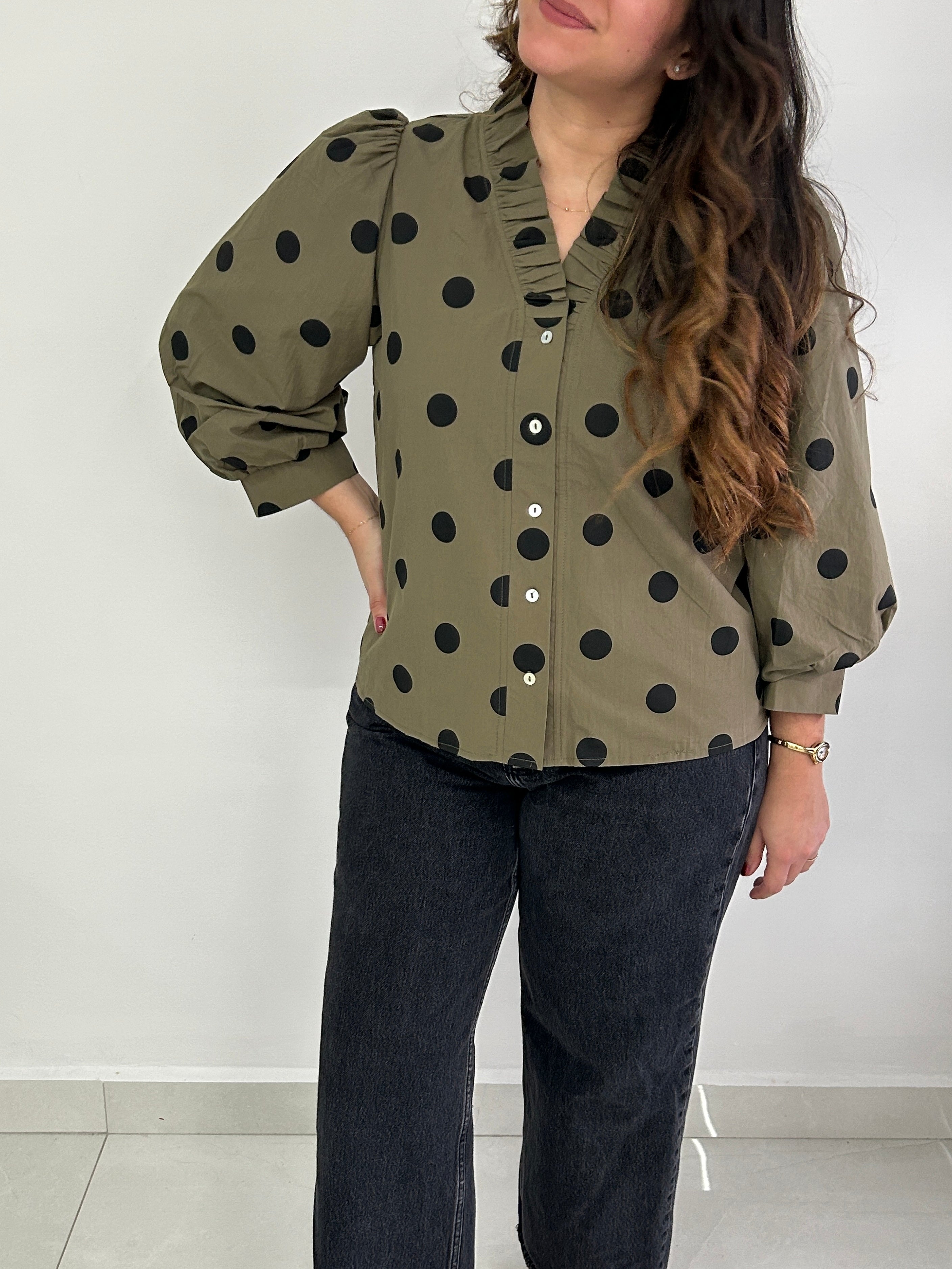 A woman wearing an olive green long-sleeve blouse with black polka dots, paired with dark jeans, standing against a plain light gray wall.