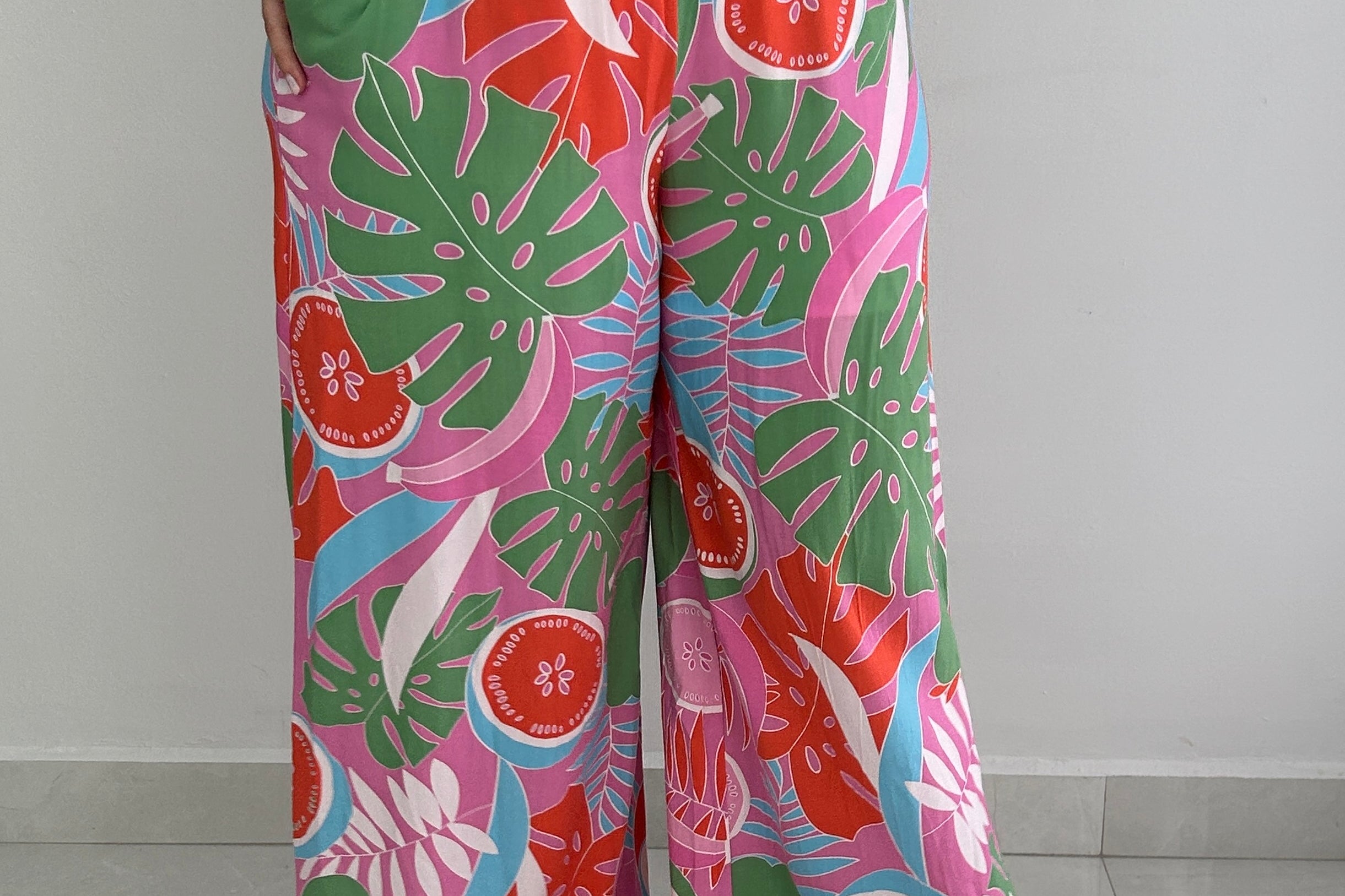 A person wearing wide-leg tropical print pants with a matching top, standing against a plain light gray wall and tiled floor. The fabric has a bold multicolor pattern with pink, green, red, and blue botanical and fruit motifs.