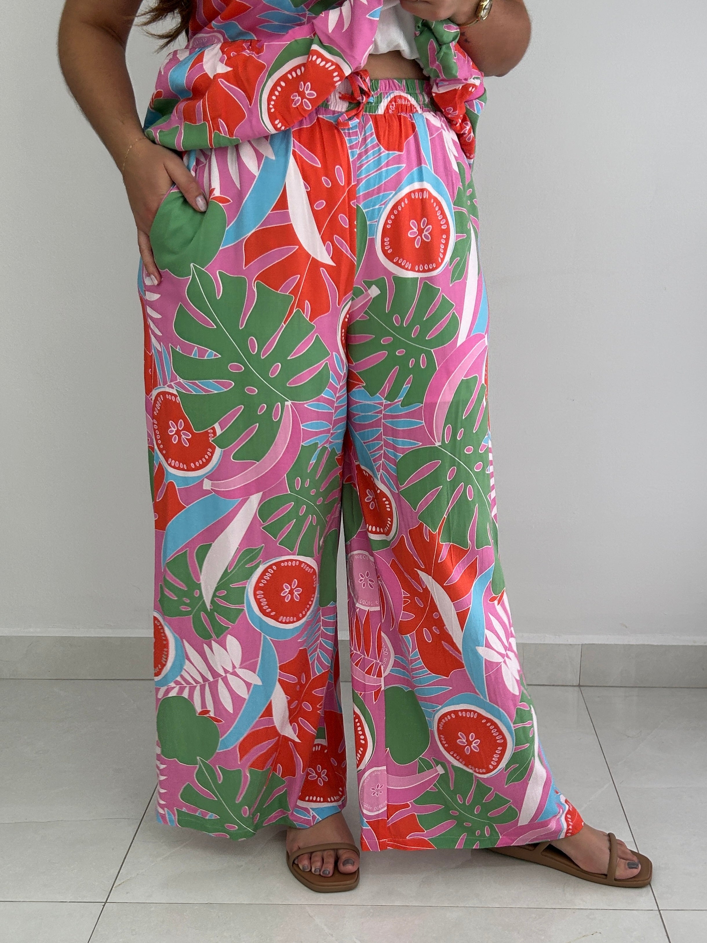 A person wearing wide-leg tropical print pants with a matching top, standing against a plain light gray wall and tiled floor. The fabric has a bold multicolor pattern with pink, green, red, and blue botanical and fruit motifs.