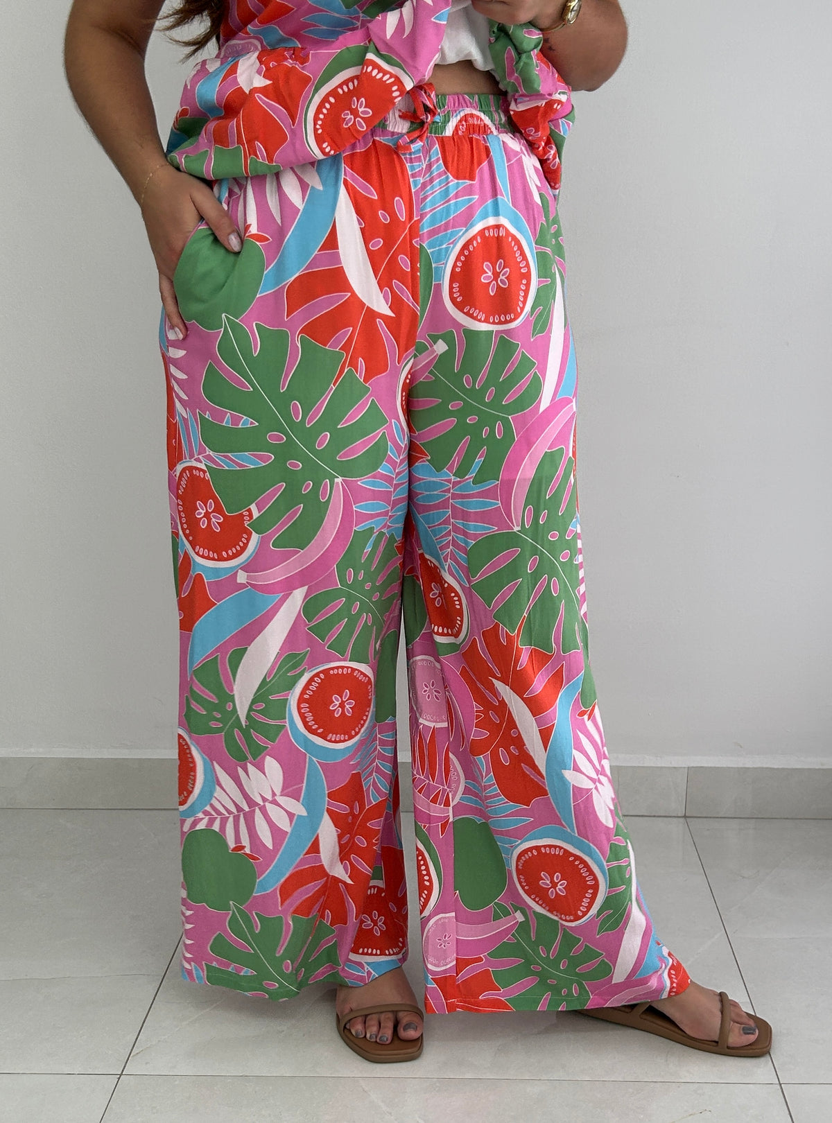 A person wearing wide-leg tropical print pants with a matching top, standing against a plain light gray wall and tiled floor. The fabric has a bold multicolor pattern with pink, green, red, and blue botanical and fruit motifs.