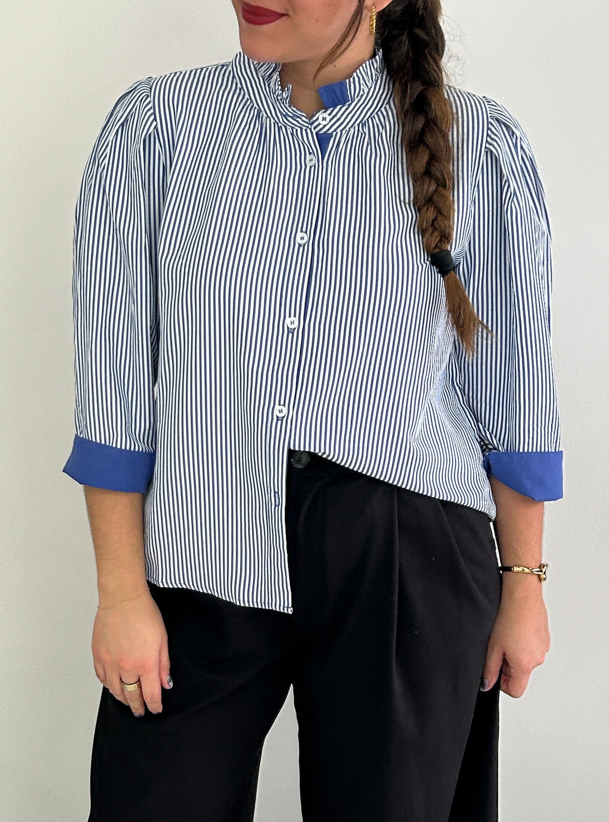 A woman wearing a blue and white vertically striped button-up shirt with solid blue contrast cuffs and a high collar, paired with black wide-leg trousers, standing against a plain light gray wall.