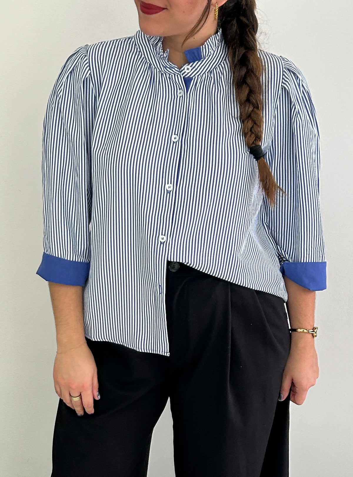 A woman wearing a blue and white vertically striped button-up shirt with solid blue contrast cuffs and a high collar, paired with black wide-leg trousers, standing against a plain light gray wall.