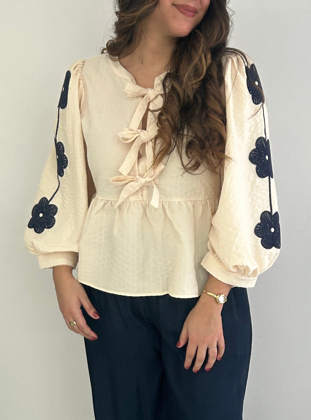 A woman modeling a cream-colored blouse with long puff sleeves, tie details at the neckline, and black embroidered flowers on the sleeves, paired with dark navy wide-leg pants and white shoes.