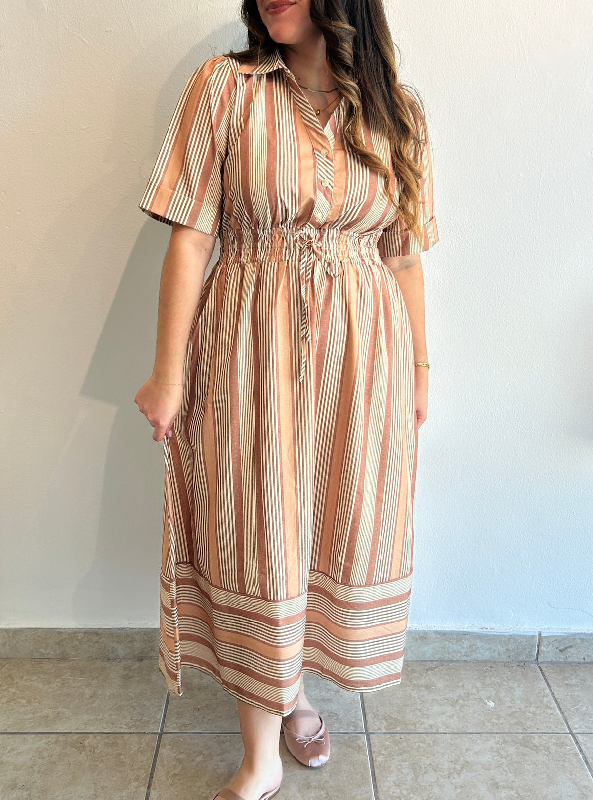 A woman wearing a short-sleeve midi dress with vertical stripes in beige, brown, and cream, standing against a plain light wall.