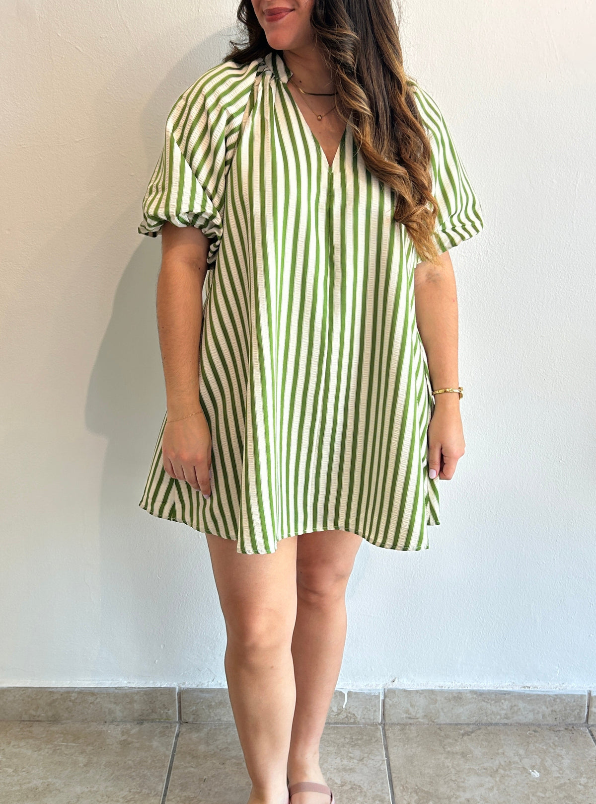 A woman wearing a short-sleeve V-neck dress with vertical green and white stripes, standing against a plain off-white wall.