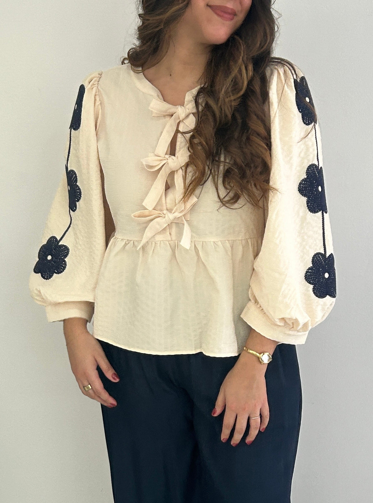 A woman modeling a cream-colored blouse with long puff sleeves, tie details at the neckline, and black embroidered flowers on the sleeves, paired with dark navy wide-leg pants and white shoes.