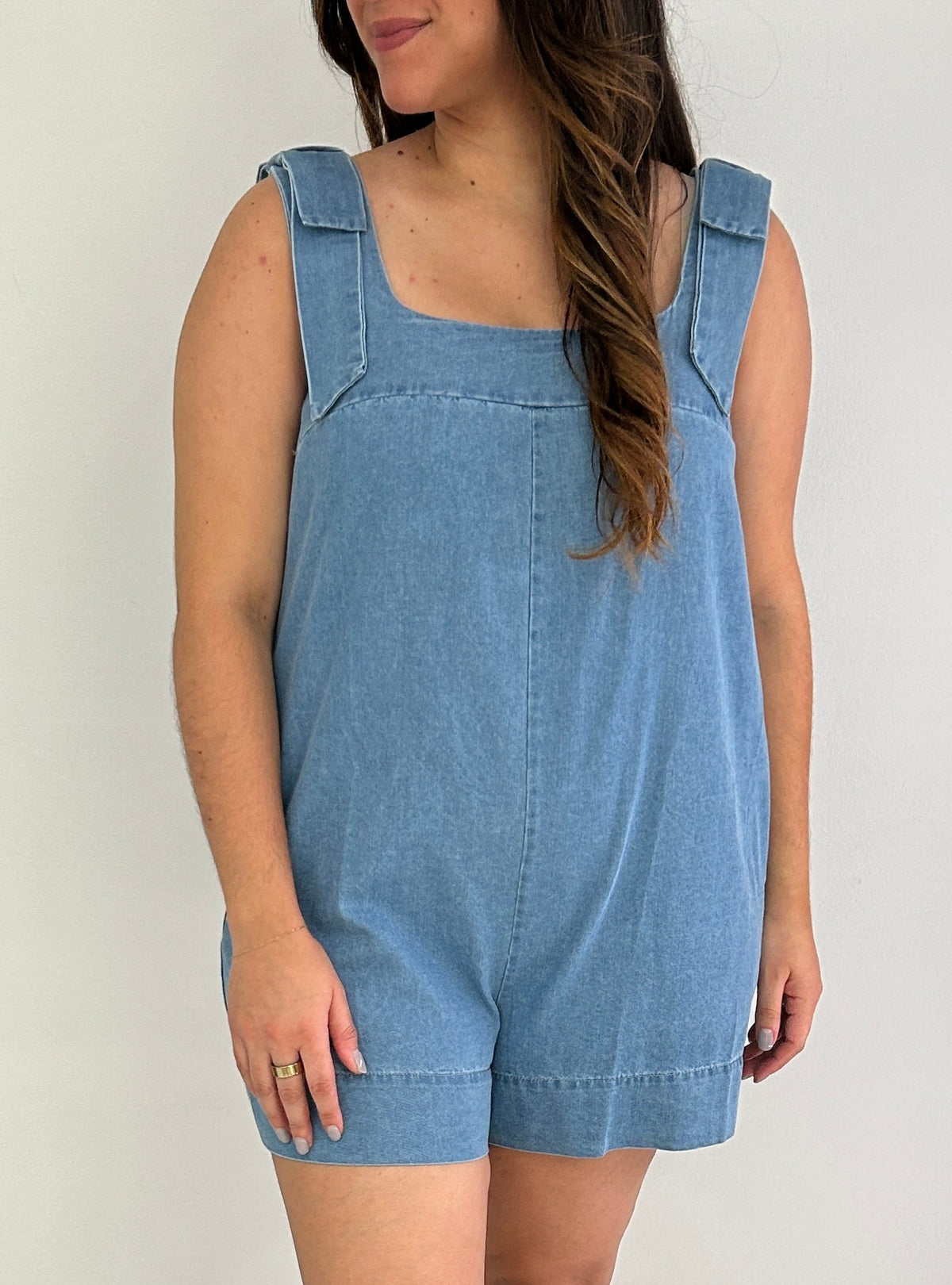 A woman modeling a light blue denim overall romper with shoulder straps, paired with pink lace-up shoes, standing against a plain white wall.