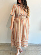 A woman wearing a short-sleeve midi dress with vertical stripes in beige, brown, and cream, standing against a plain light wall.