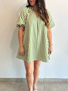 A woman wearing a short-sleeve V-neck dress with vertical green and white stripes, standing against a plain off-white wall.